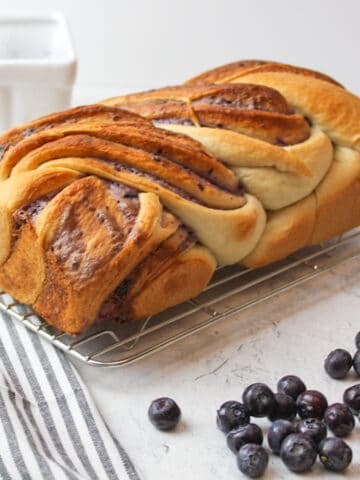 a loaf of blueberry cream cheese babka on a wire rack with fresh blueberries to the side.