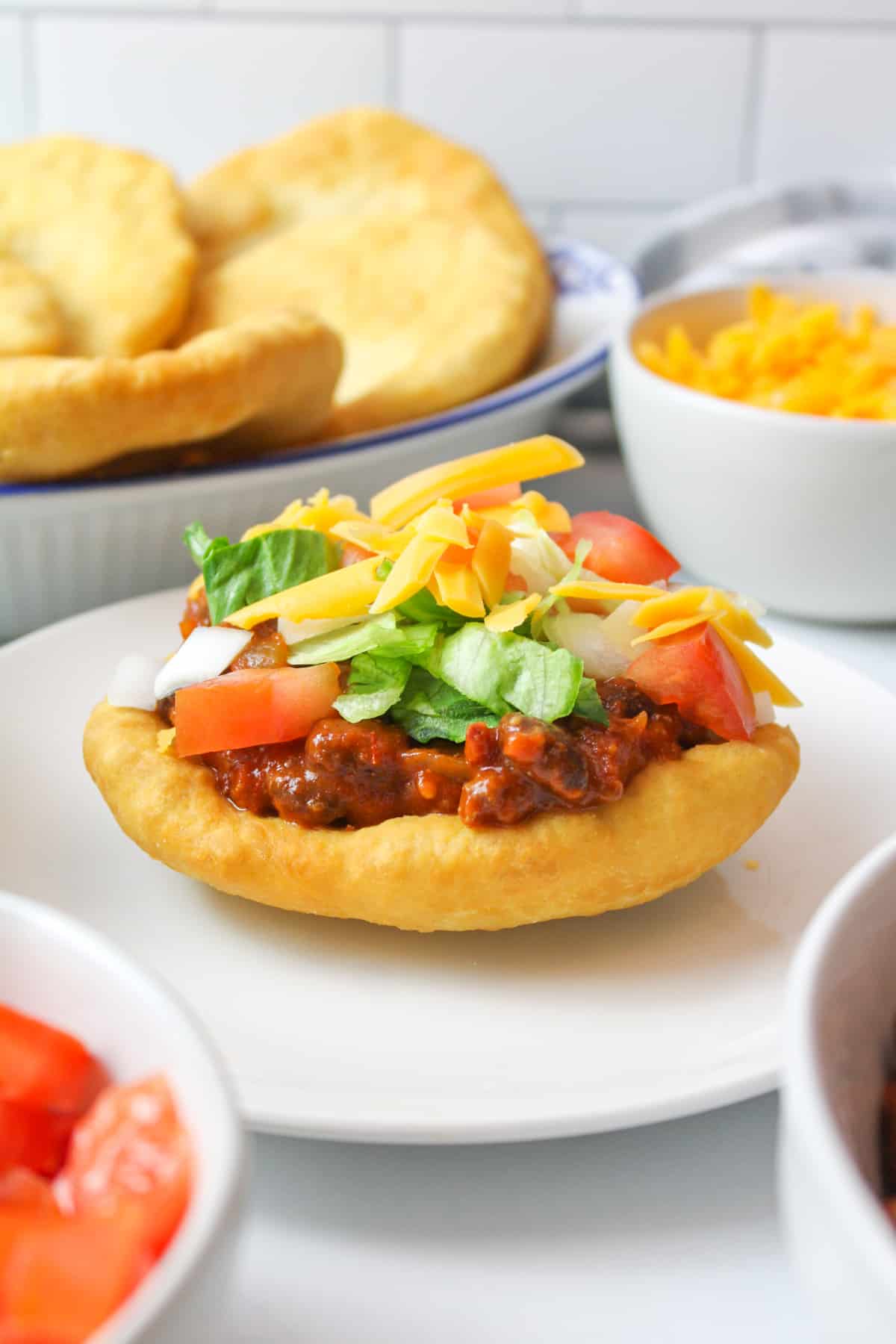 a piece of fry bread on a plate topped with chili, lettuce, tomatoes, onion and cheese to make an indian taco. More frybread and toppings in background