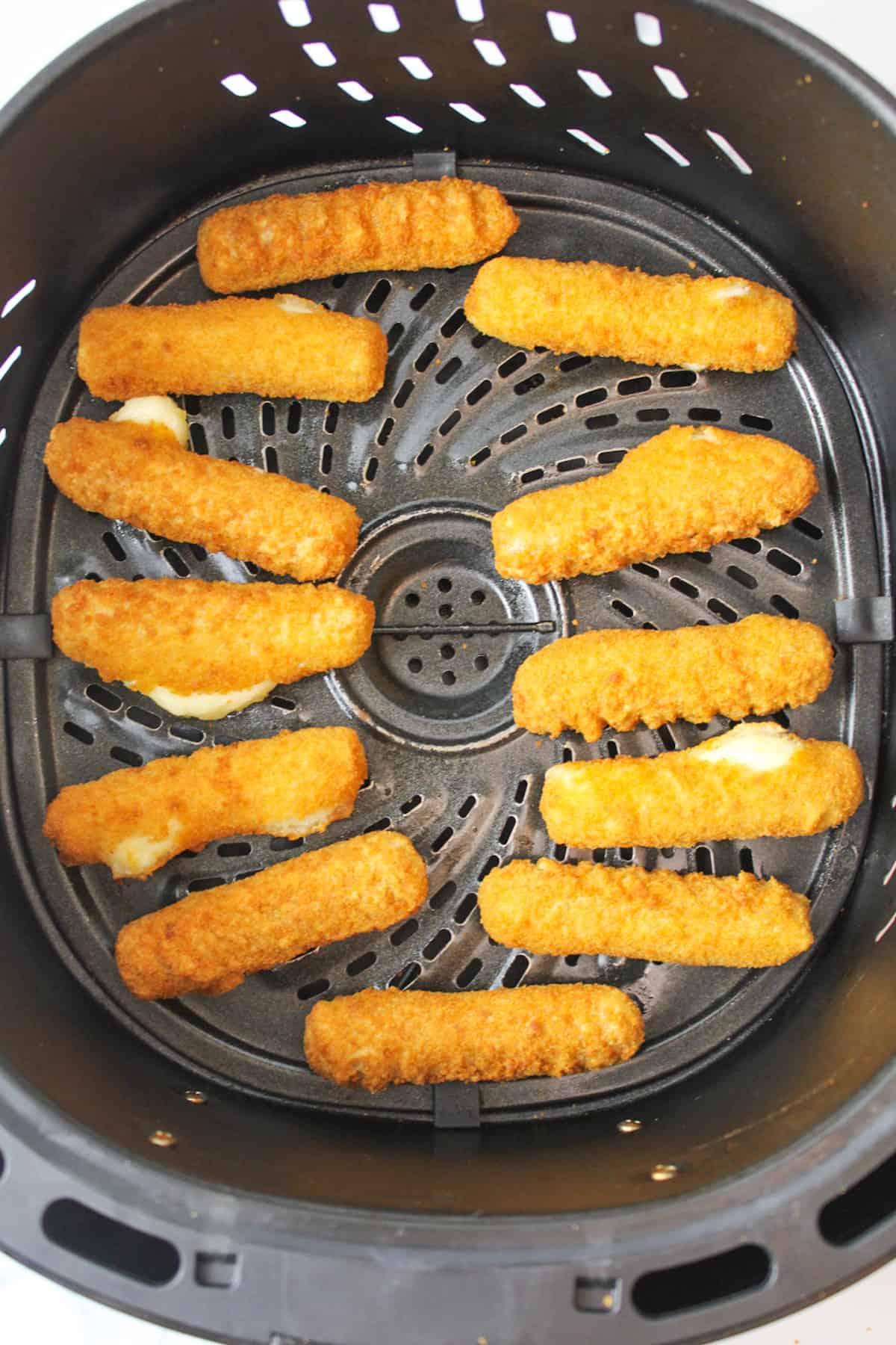 cooked mozarella sticks in an air fryer basket, some having cheese pop out