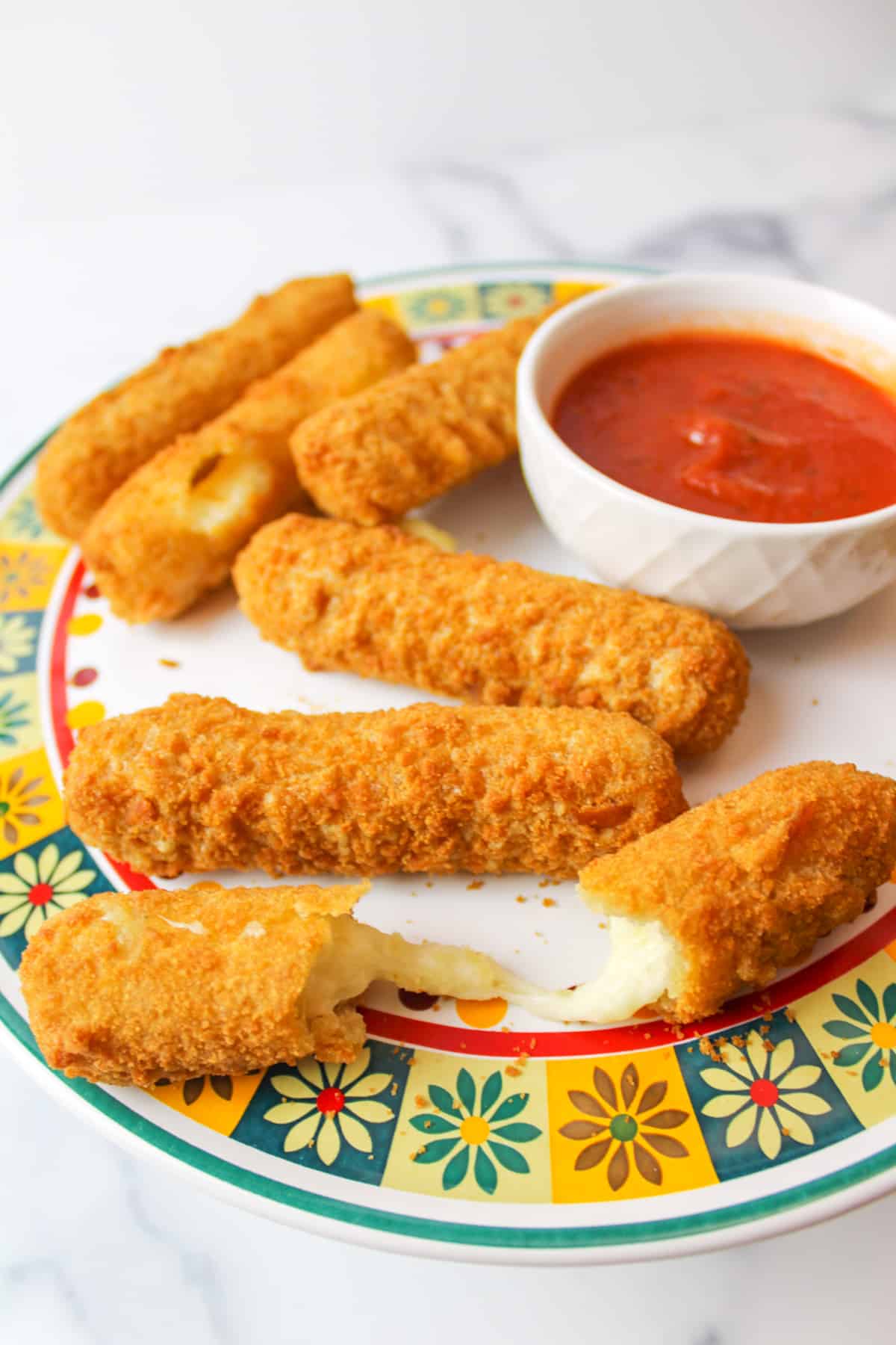 a plate of mozzarella sticks next to a bowl of sauce one mozzarella stick is broken in half to reveal stringy cheese inside