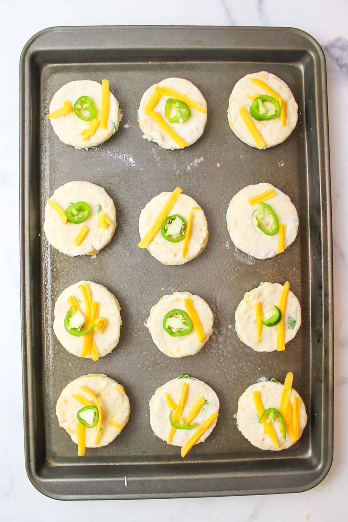 Jalapeno cheddar biscuits topped with shredded cheese and sliced jalapenos on a baking sheet.
