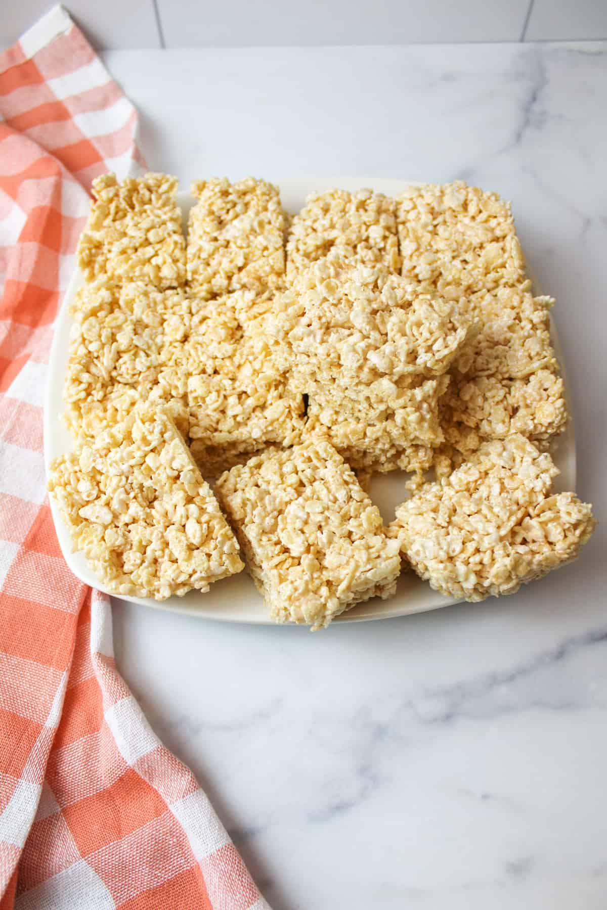 Rice Krispies treats cut into squares on plate, pink checkered towel beside.