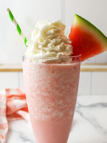 a glass of watermelon milkshake topped with whipped cream and a slice of watermelon.