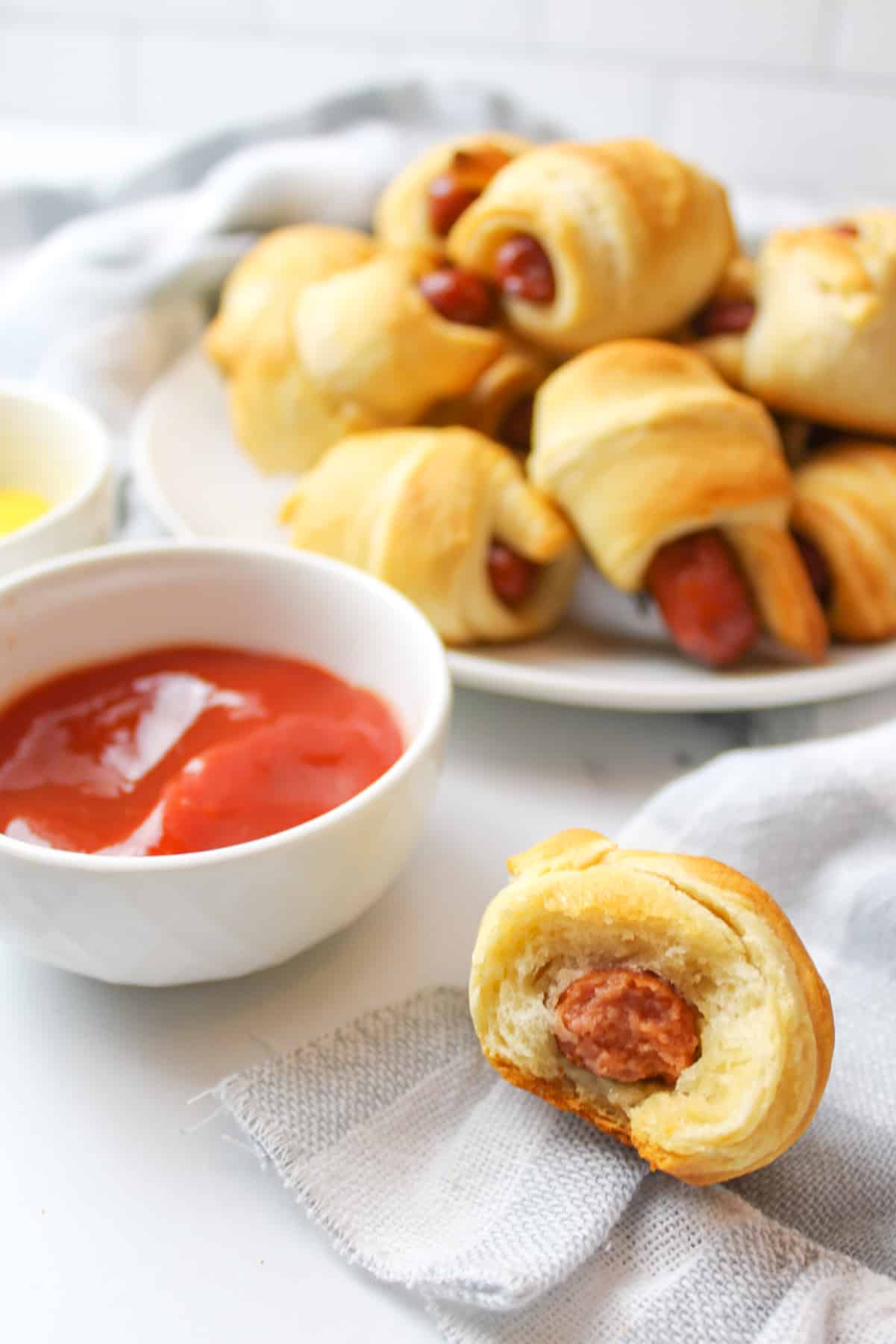a mini pig in a blanket that had a bite removed with more piggies on a plate in background next to bowls of condiments