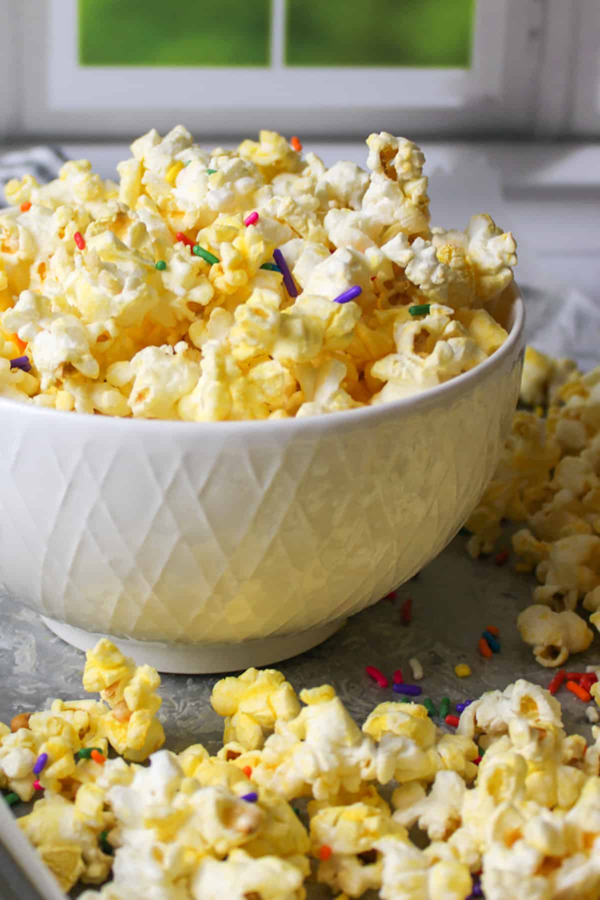 Almond bark popcorn with sprinkles in a bowl and more on a surrounding baking sheet.