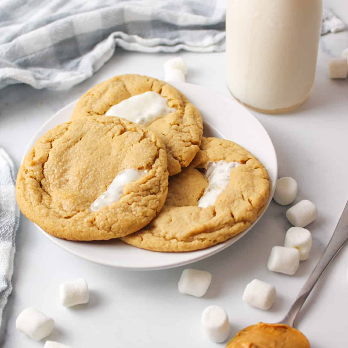 three peanut butter marshmallow cookies on a plate with milk, mini marshmallows and peanut butter off to the sides.