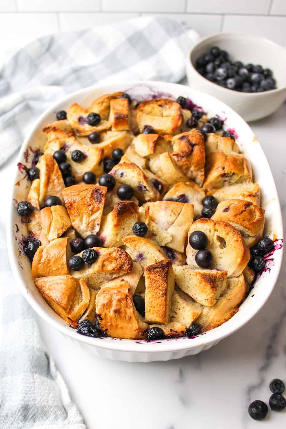 Blueberry bagel bread pudding topped with blueberries, with a bowl in the background.