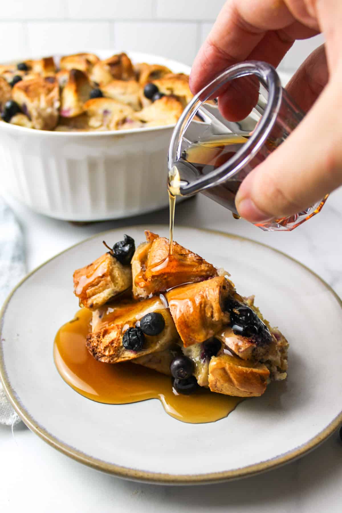 a hand drizzling syrup over the top of a scoop of blueberry bagel bread pudding on a plate with more bread pudding in the background