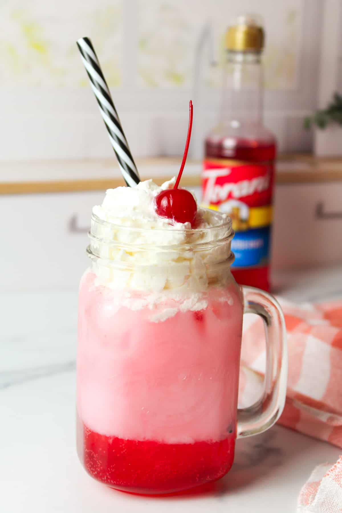 a glass full of italian soda topped with whipped cream and a cherry with a bottle of strawberry coffee syrup in the background