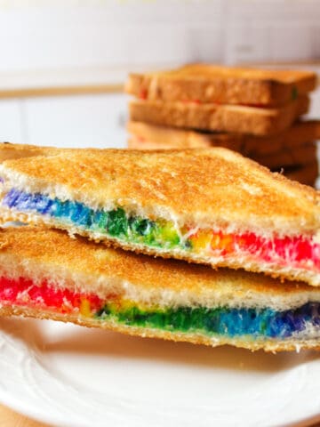 sliced rainbow grilled cheese on a white plate with more sandwiches in background.