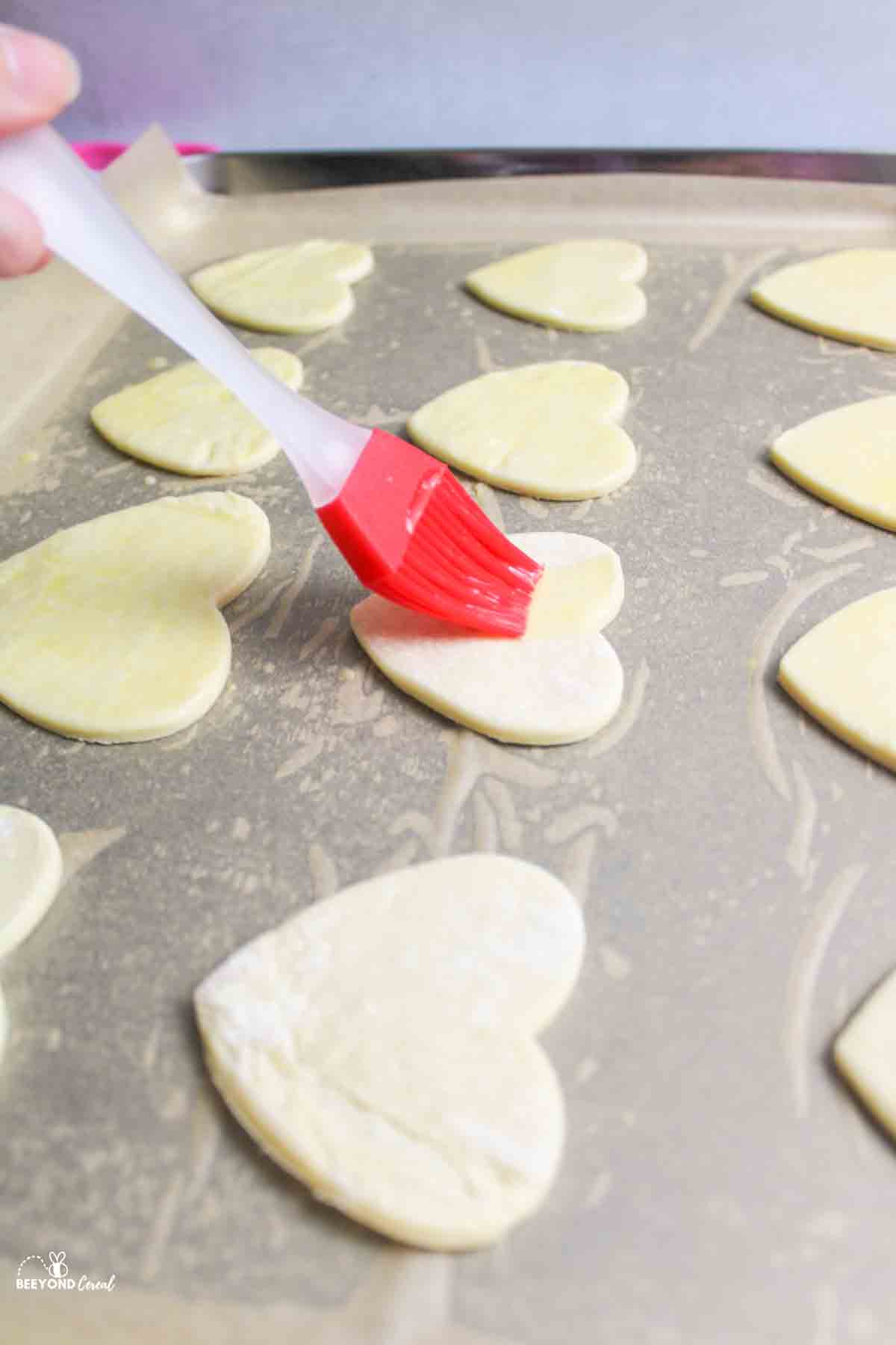 brushing eggwash over puff pastry hearts on a baking sheet