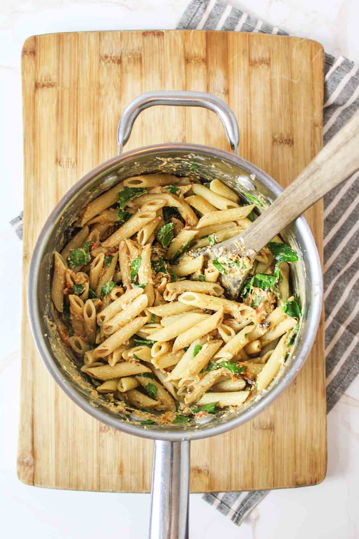 a wooden spoon stirring cream cheese mixture and spinach into cooked pasta in pot.