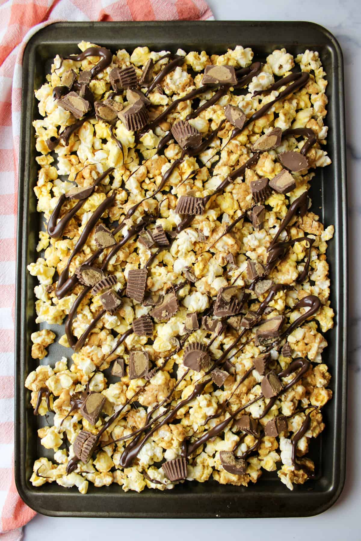 chocolate drizzled over peanut butter coated popcorn on a baking sheet and topped with chunks of peanut butter cups