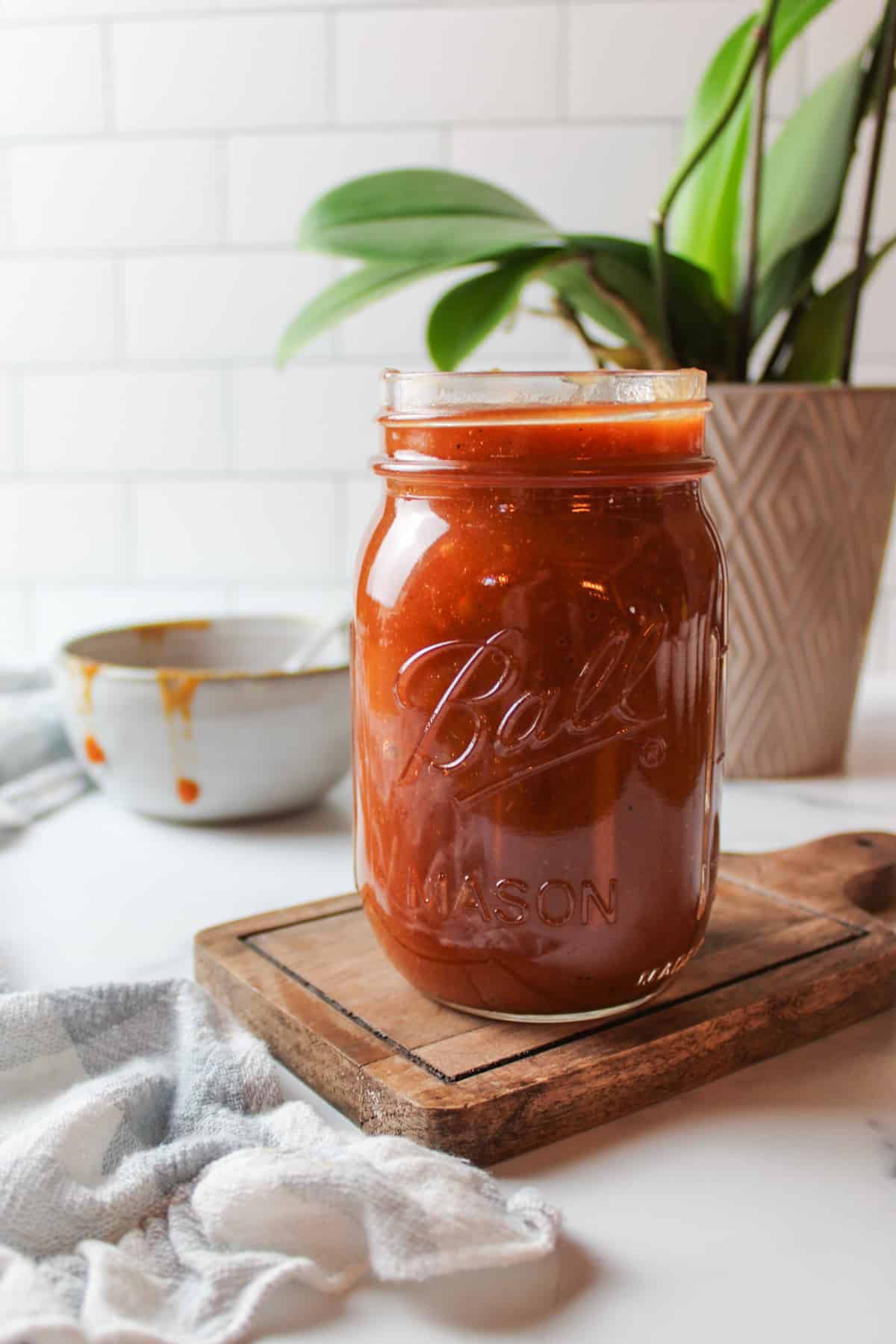 a jar filled with homemade bbq sauce and a messy bowl of sauce in the background next to a green leaf plant