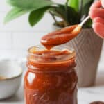 a hand holding a spoon of sauce above a jar filled with barbecue sauce