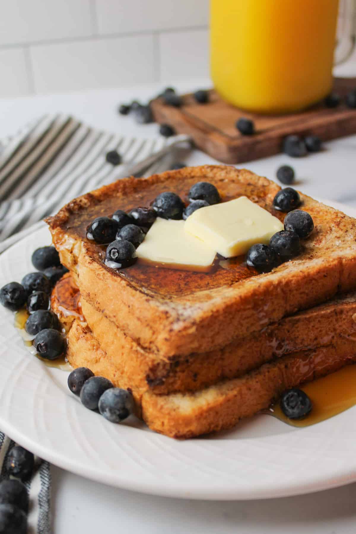 Stacked French toast on plate with blueberries, butter slices, and maple syrup.