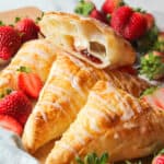 close up of strawberry cream cheese turnovers with fresh berries and one turnover sliced in half stacked on glazed full turnovers