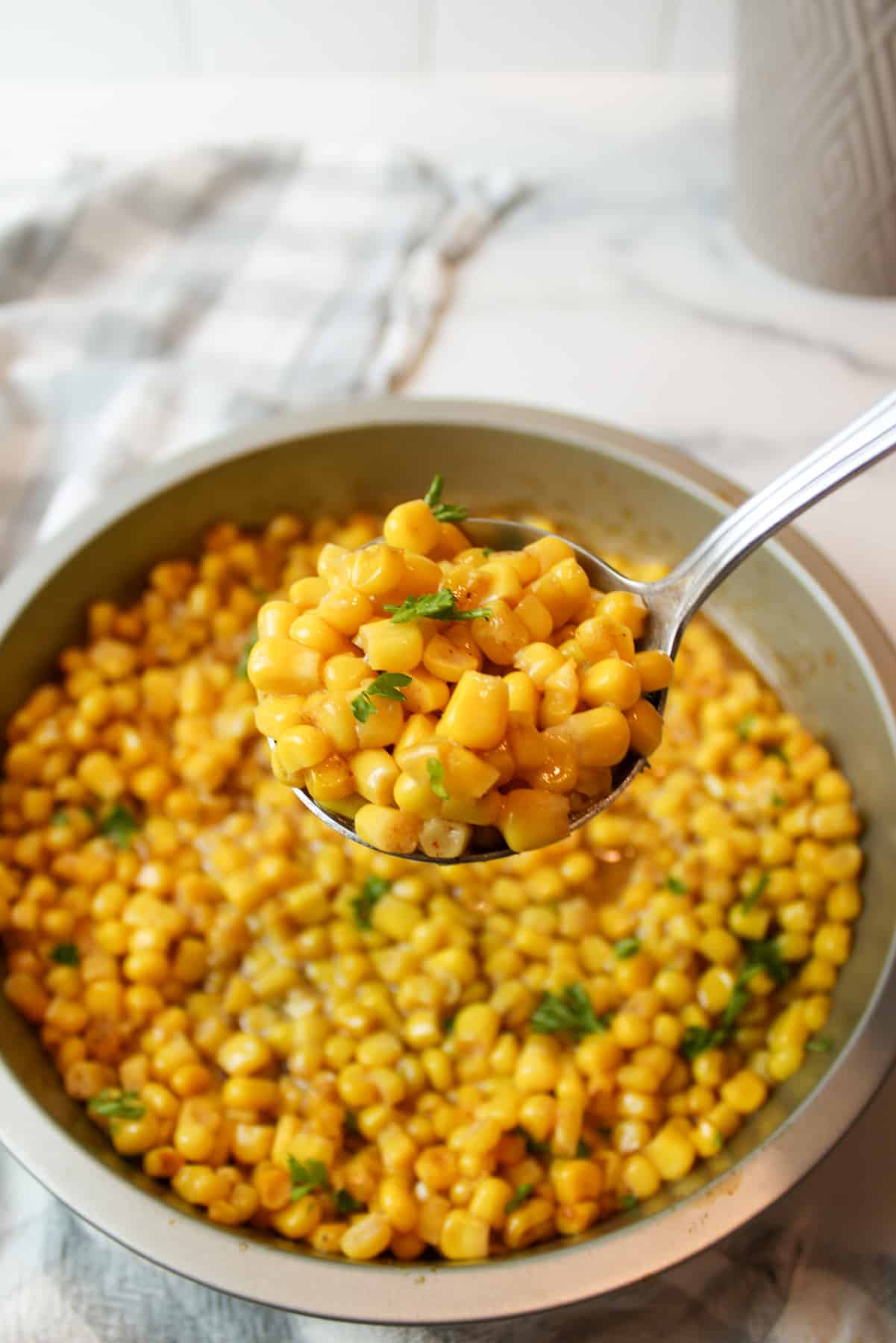 a spoon holding up a scoop of cooked corn kernels and garnished with fresh parsley.