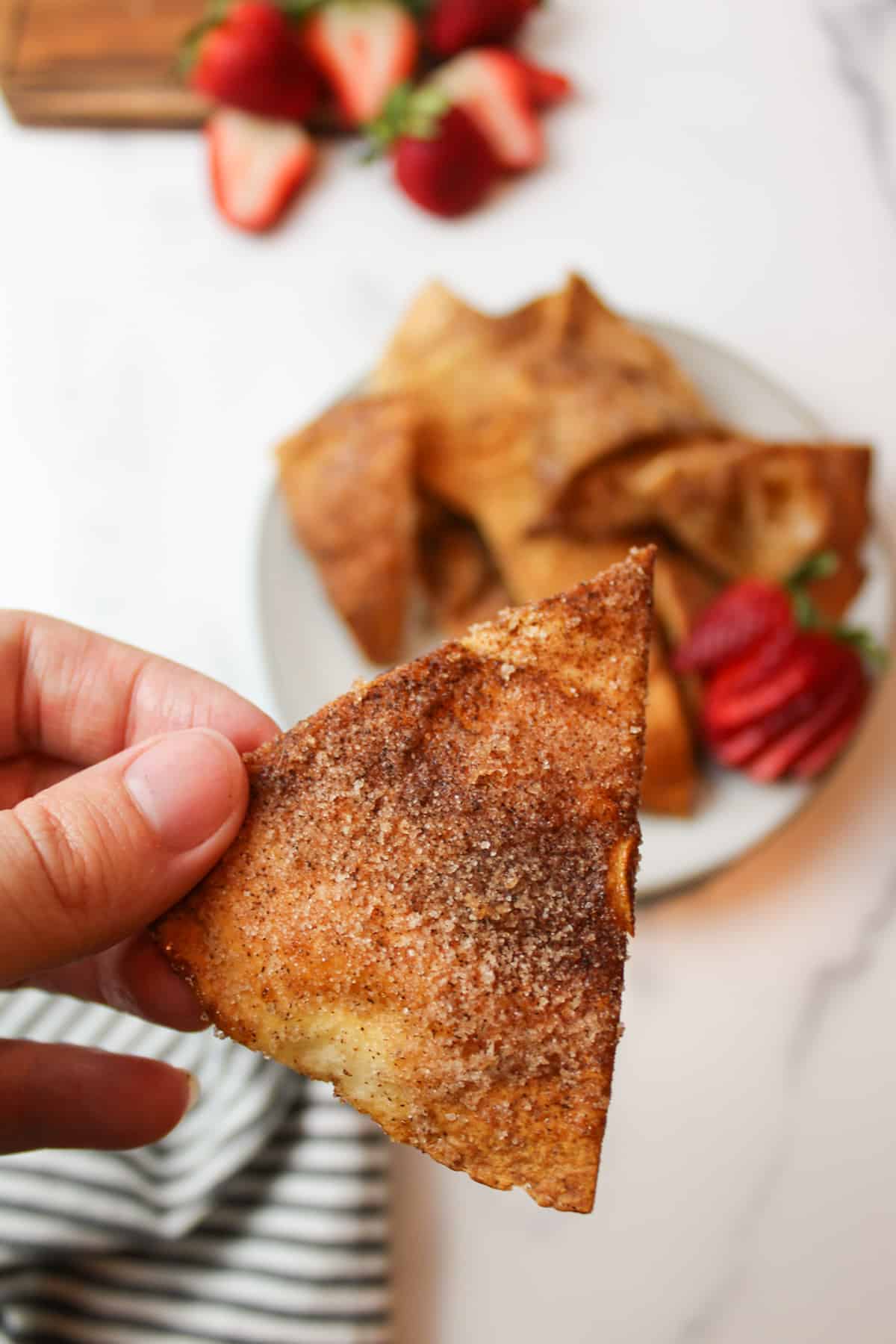 a hand holding up a crispy cinnamon sugar tortilla chip with a plate holding more in the background next to fresh strawberries.
