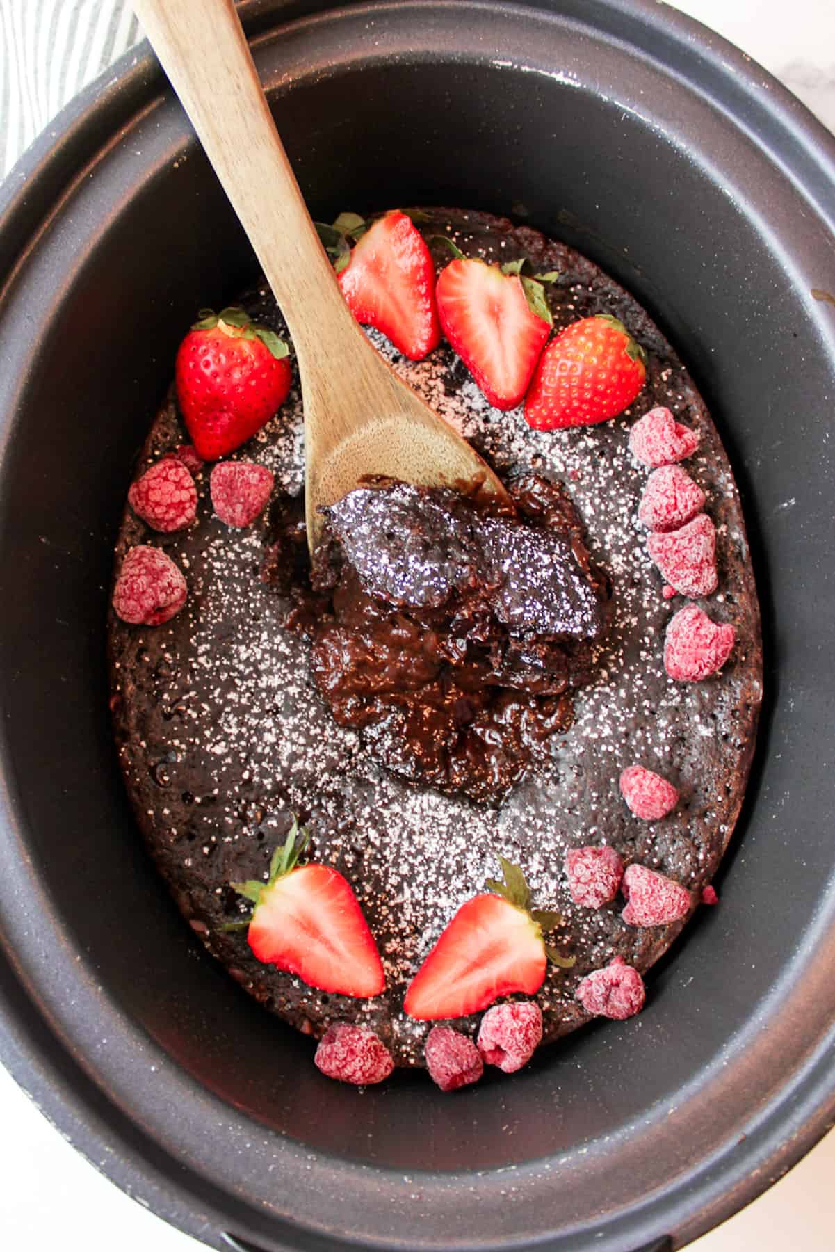 aerial view of crockpot filled with chocolate lava cake topped with powdered sugar and mixed red berries with a wooden spoon disturbing the center to revel gooey chocolate