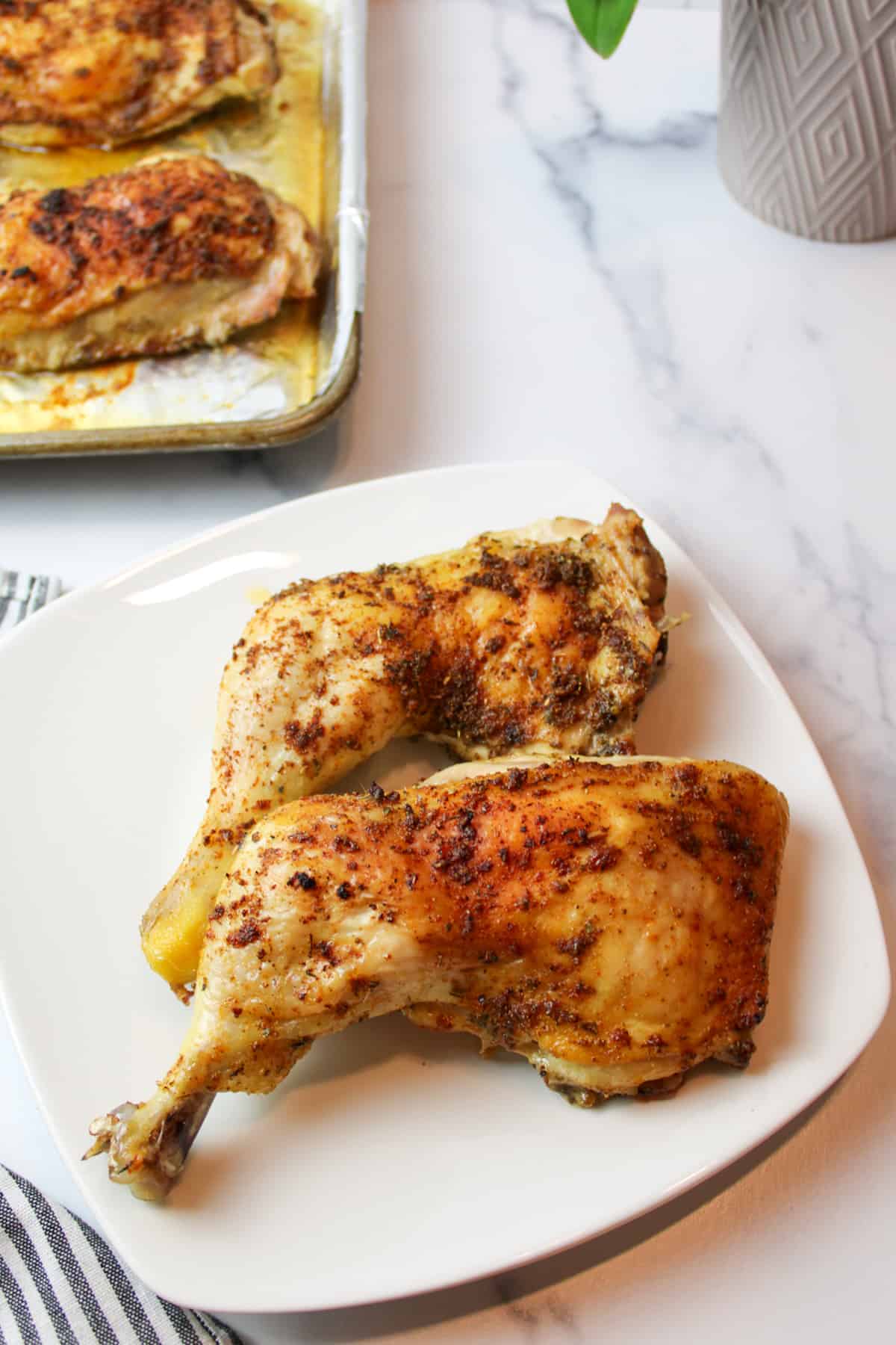two crispy baked chicken quarters on a plate with more on a baking sheet in background