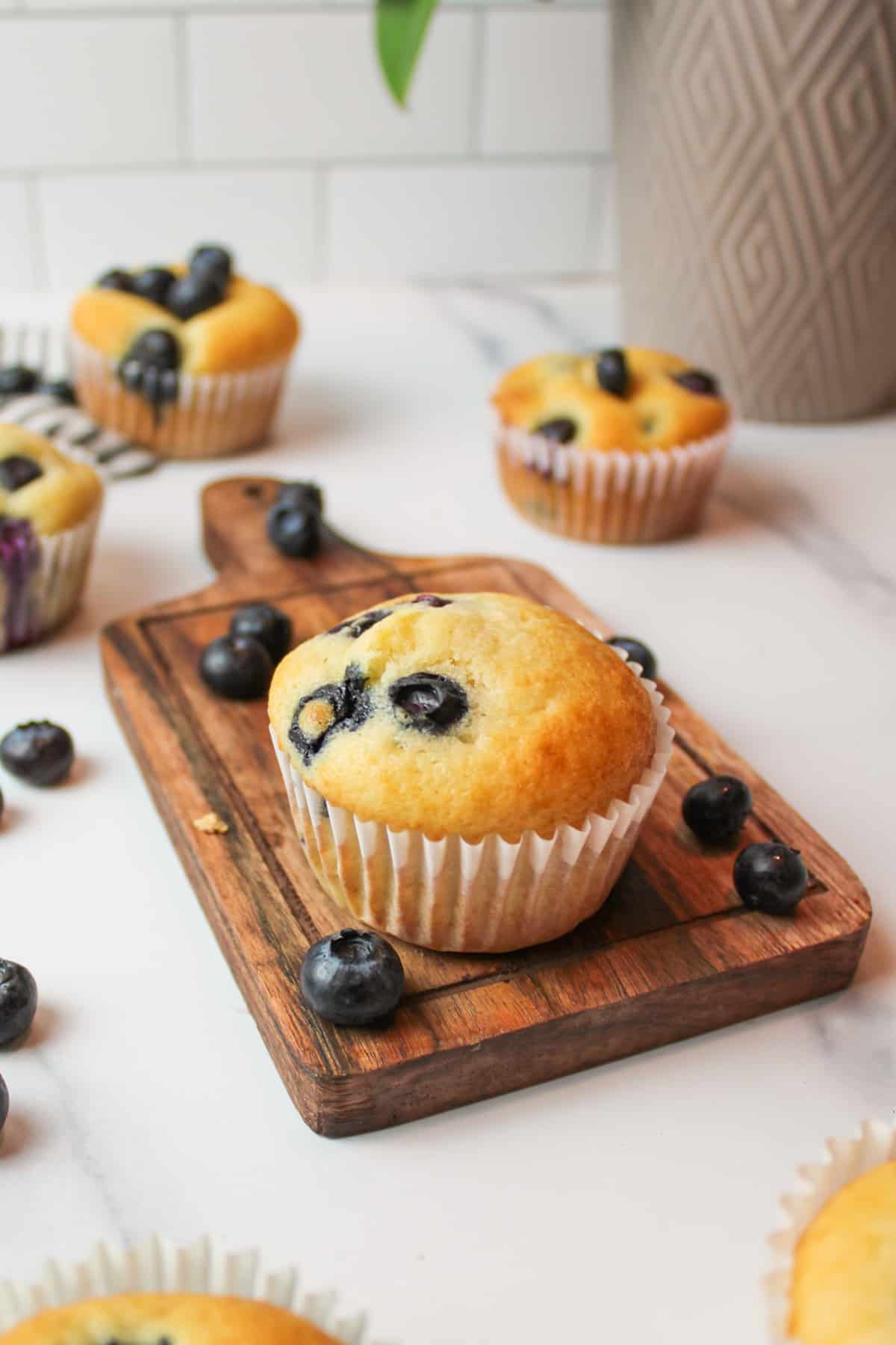 blueberry muffins and scattered blueberries with a muffin in the center of the frame on a wooden cutting board