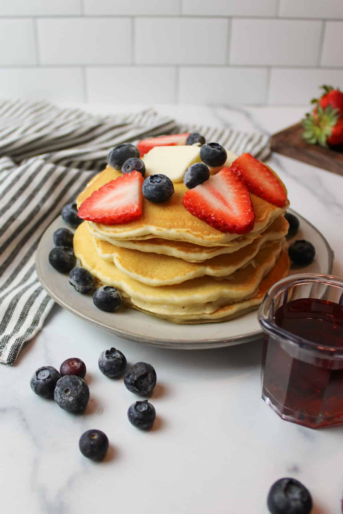 a stack of fluffy pancakes topped with fresh berries and butter with maple syrup in a container to the side.