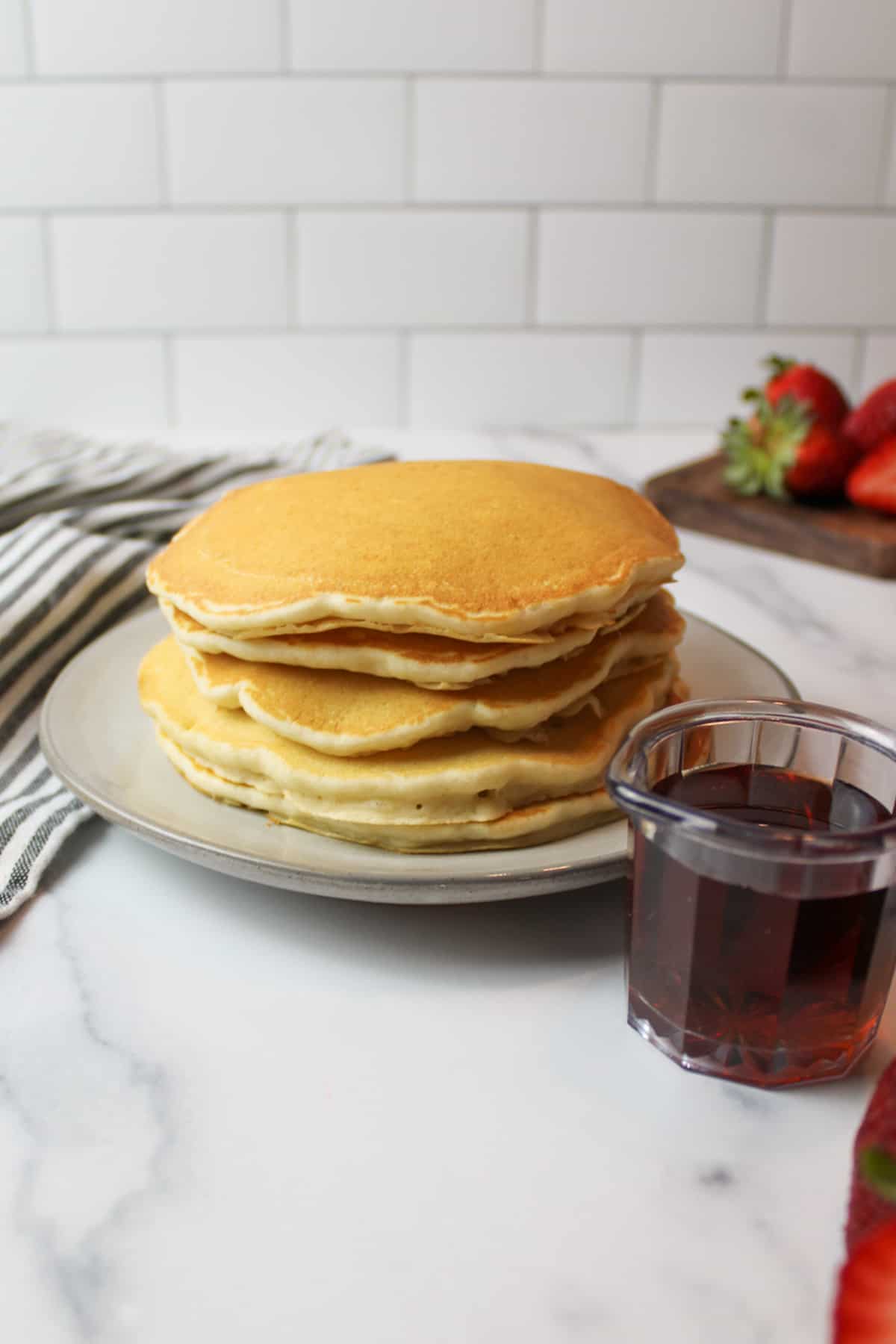 a stack of pancakes on a plate with a cup of maple syrup to the side.