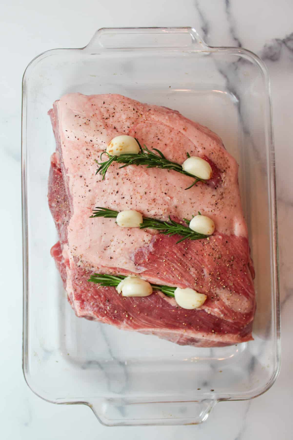 fresh rosemary and garlic cloves tucked into three slits cut into a pork shoulder roast in a baking dish