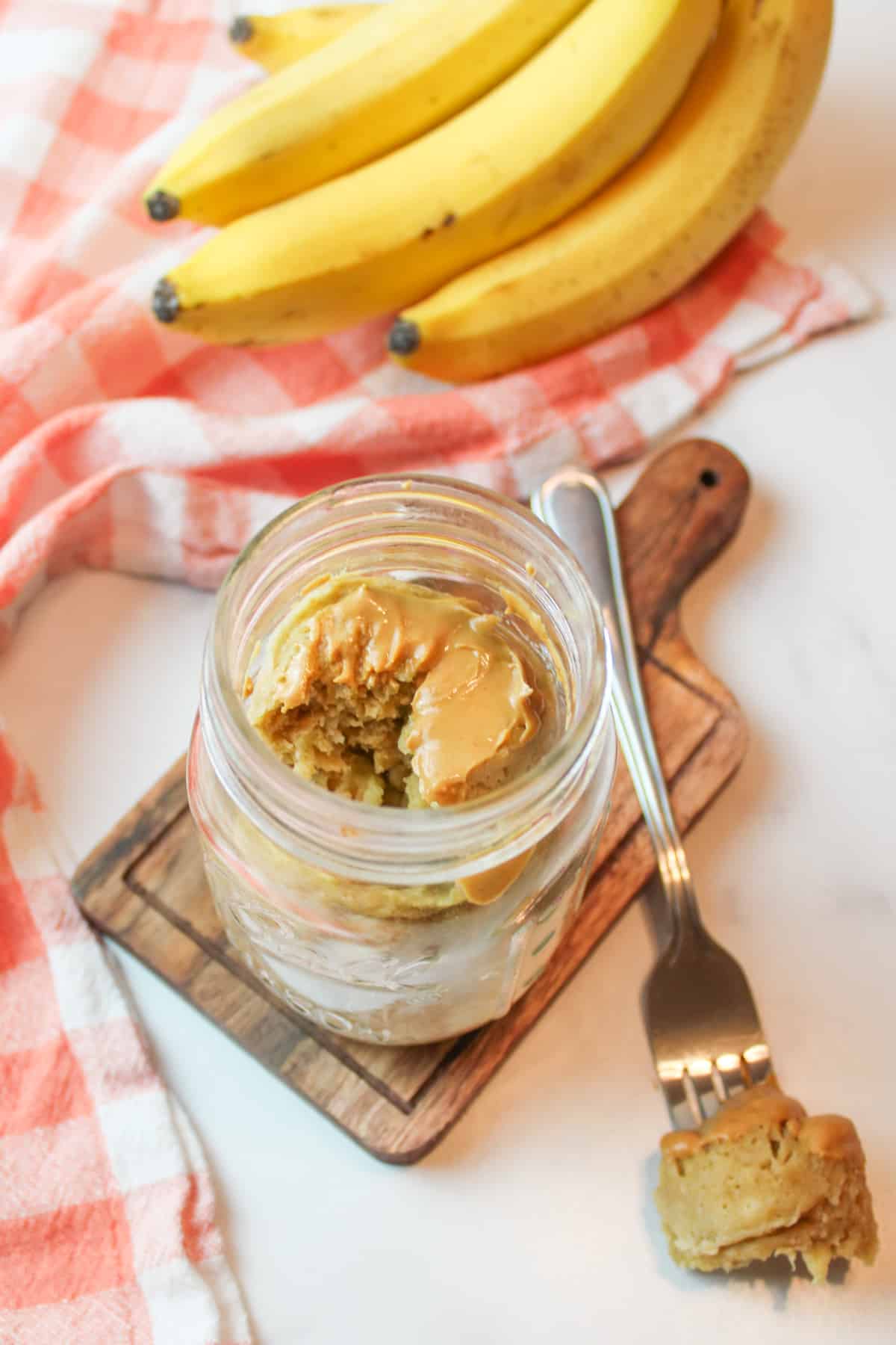 Peanut butter banana mug cake in mason jar, with fork and ripe bananas in background.