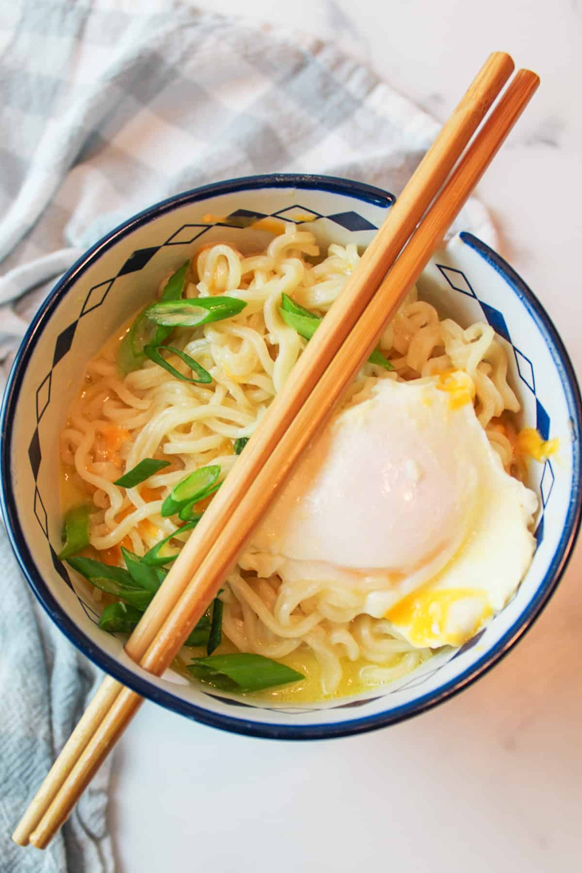 Chopsticks rest on bowl of cheesy ramen with poached egg, garnished with green onion.