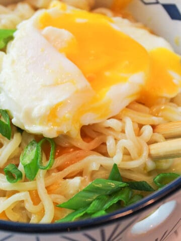 an upclose view of a bowl filled with cheesy ramen and a soft poached egg on top garnished with sliced green onions