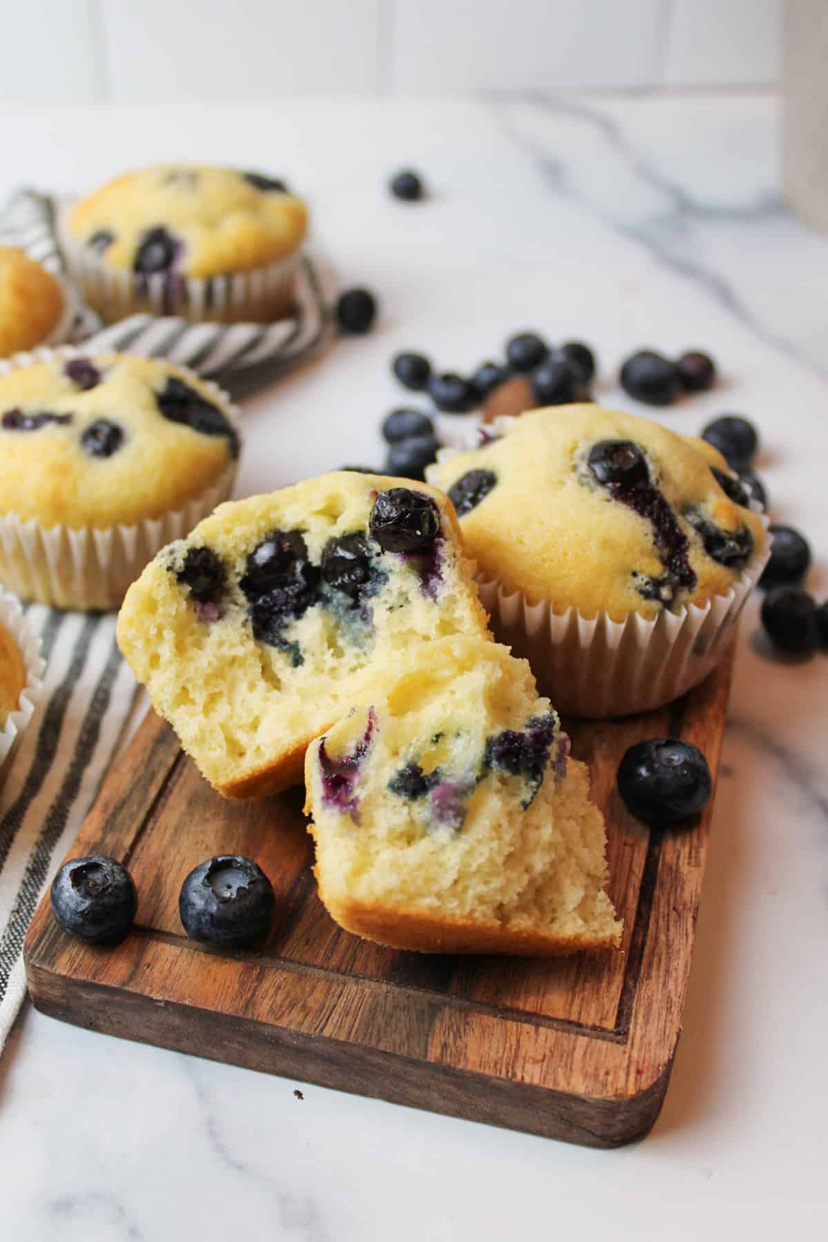 Blueberry muffins on and off wooden cutting board, scattered blueberries around.