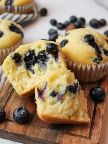 blueberry muffins on a wooden cutting board with one broken open to reveal the inside crumbs and scattered blueberries around them.
