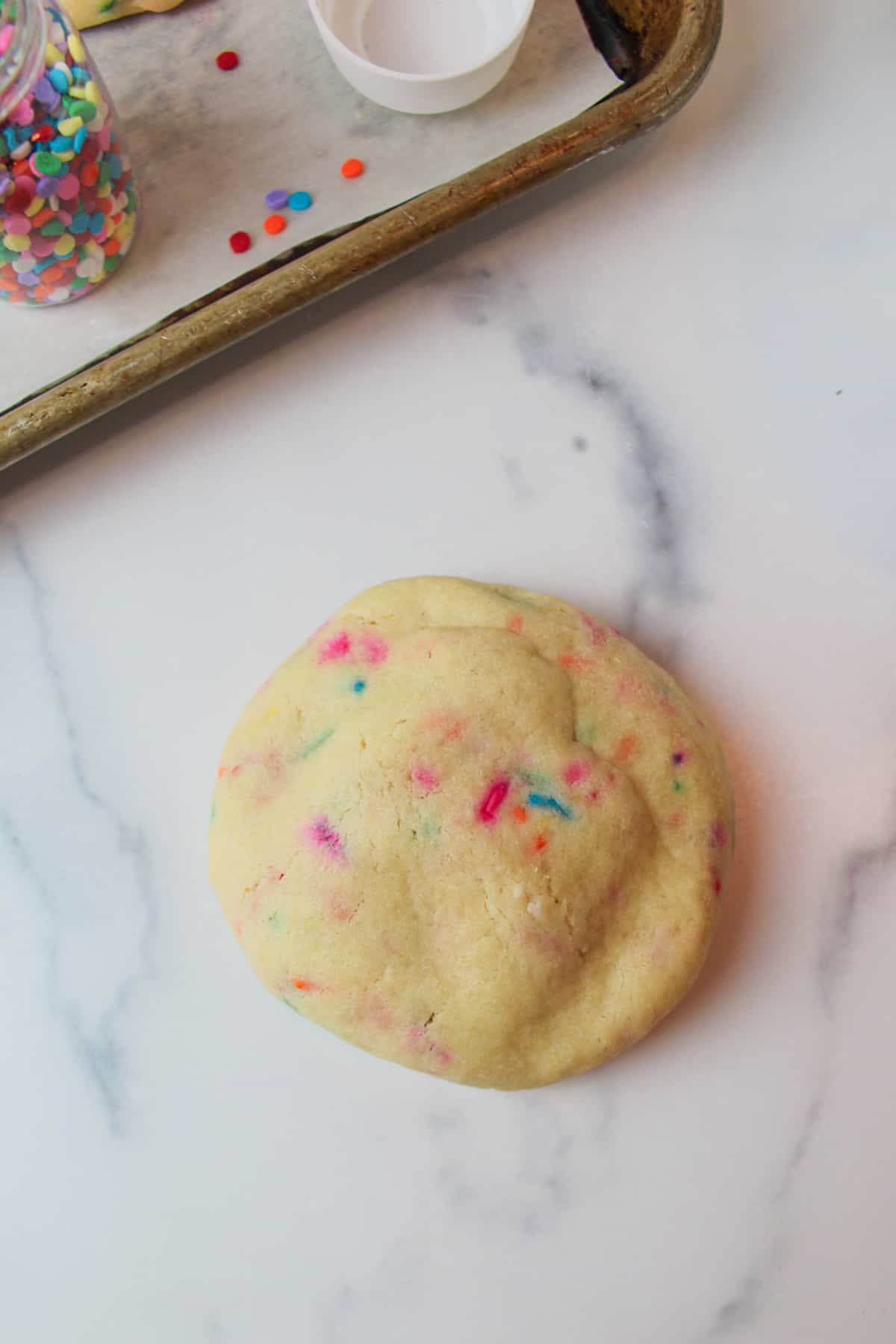 a baked birthday cake cookie next to a baking sheet with sprinkles on it