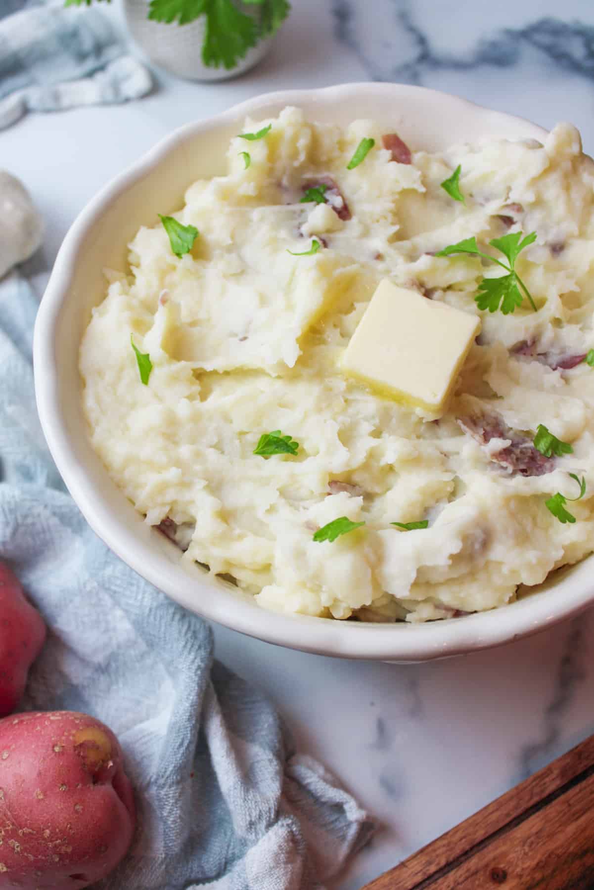 a bowl of creamy mashed potatoes with a pad of butter on top and some raw red potatoes to the side