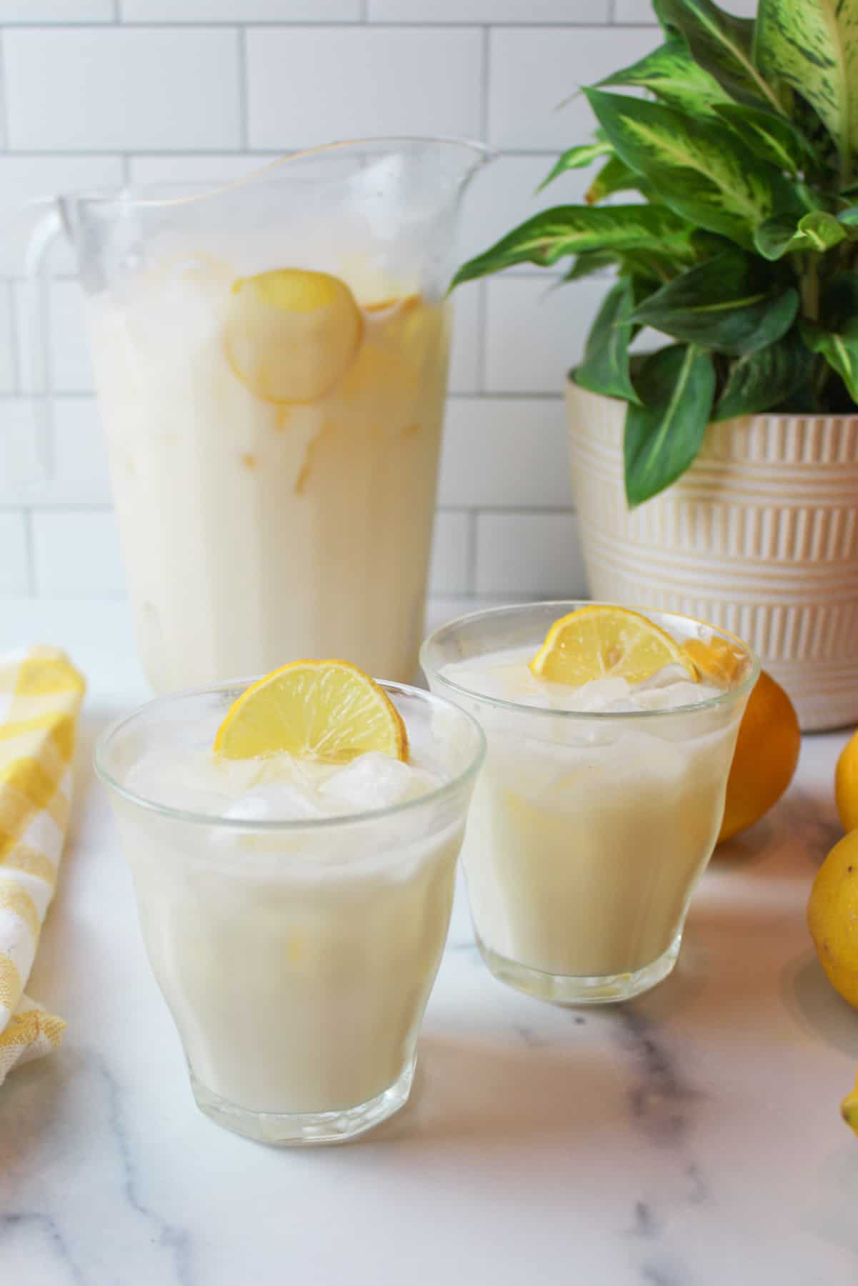 two glasses of creamy lemonade garnished with a slice of lemon and a pitcher of lemonade in the background