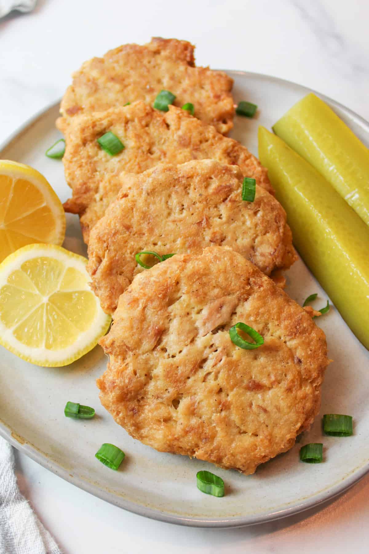 Four crispy tuna patties on plate with green onion, lemon, pickle spears.