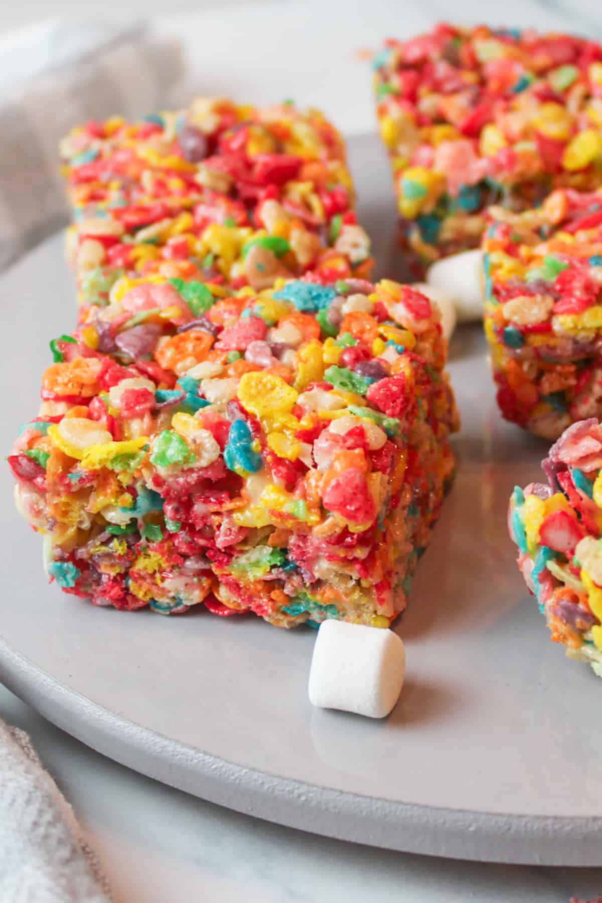 sliced squares of fruity pebbled treats on a wooden plate with a few mini marshmallows around them.