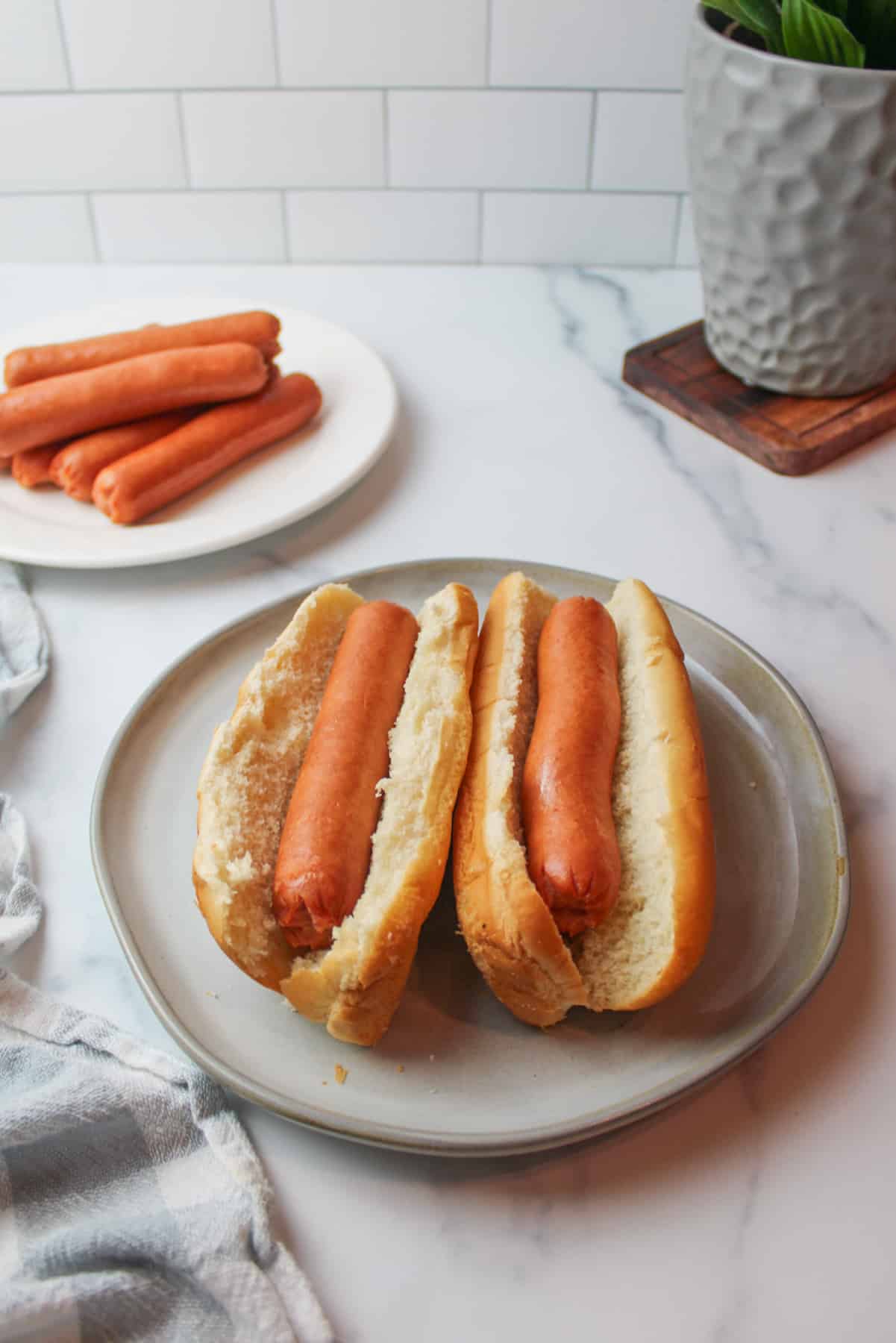 two hot dogs on buns on a plate with more cooked hot dogs on a plate in the background.