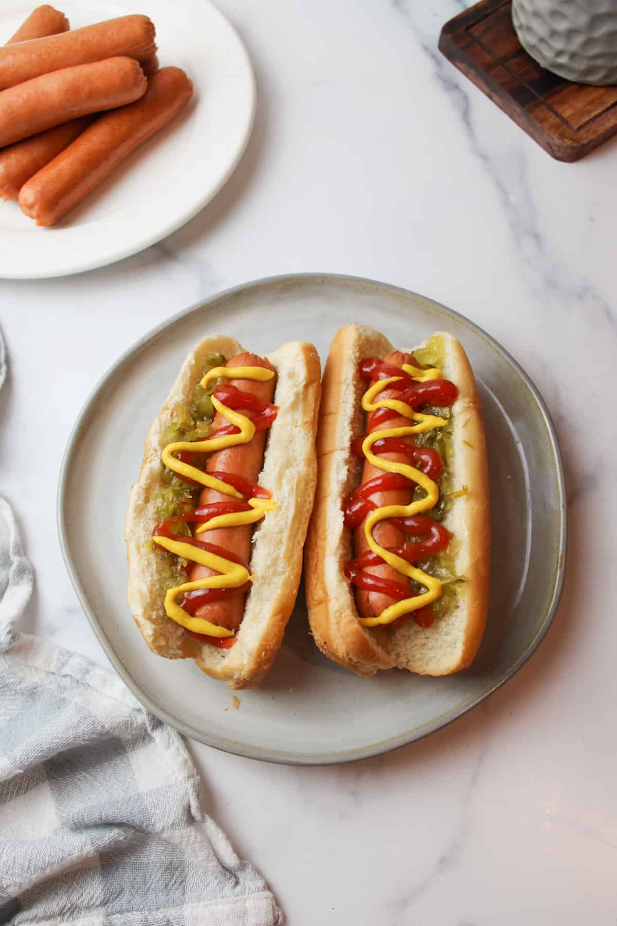 Hot dogs on plate with ketchup, mustard, relish, cooked hot dogs on side plate.