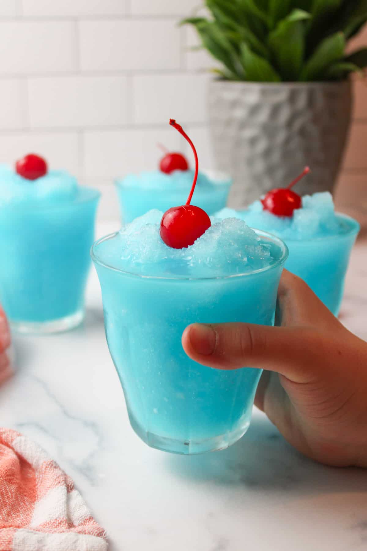 a hand holding up a glass of blue kool aid slushie with a cherry on top.