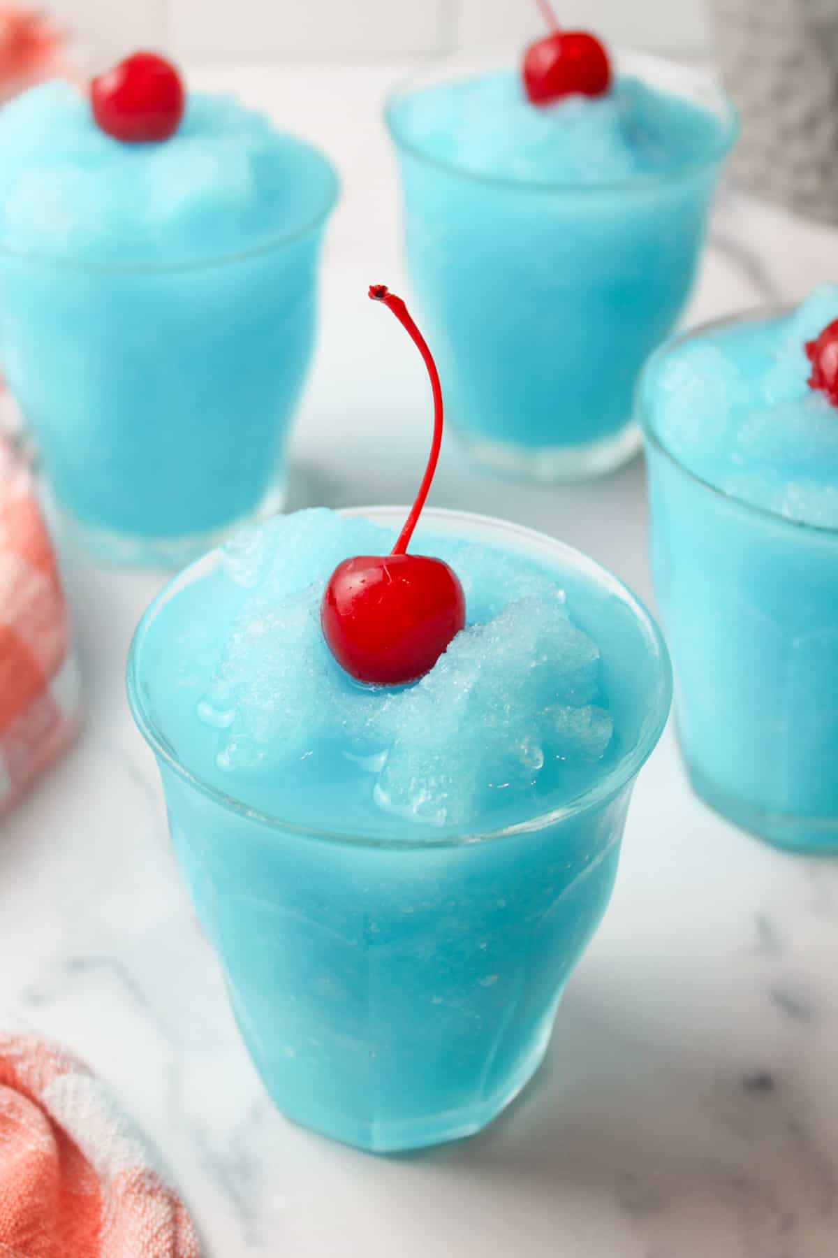angled view into a glass of blue kool aid slishie with a cherry on top and mroe drinks in background