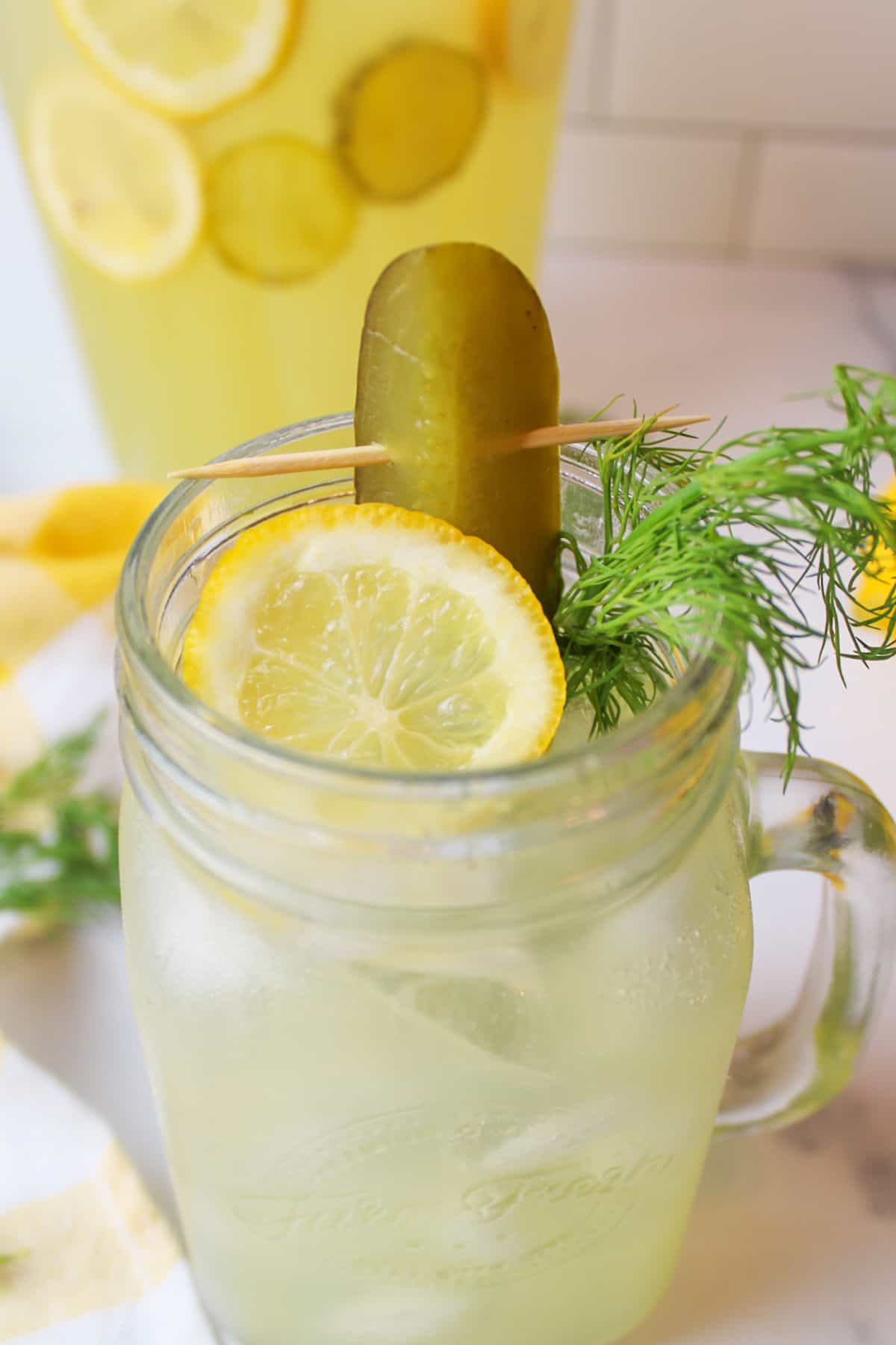 a glass of pickle lemonade garnished with lemon slice a pickle spear and dill weed.