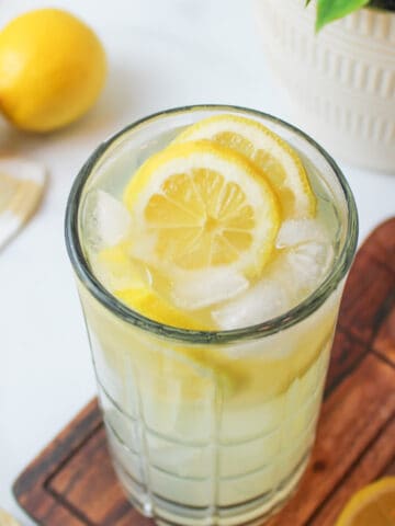 angled view into a glass of ice cold lemonade with sliced lemons on a wooden cutting board and fresh lemons to the side