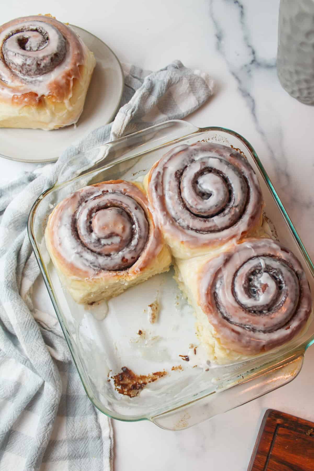 aerial view of 4 cinnamon rolls with icing, 3 of them are in a baking dish and one is on a plate to the side