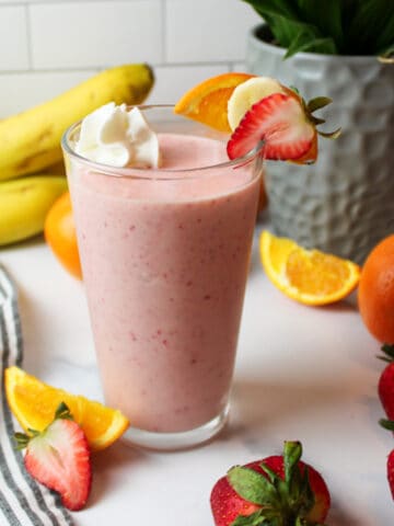 a tall glass full of strawberry orange smoothie with fresh fruits around it.