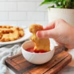 an upclose view of a child hand dipping a dino nugget into a small bowl of ketchup