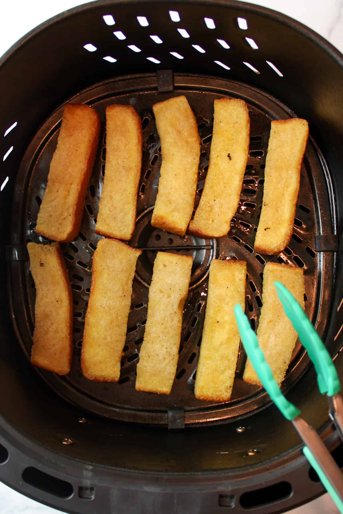 french toast sticks in an air fryer basket with a pair of tongs to the side.