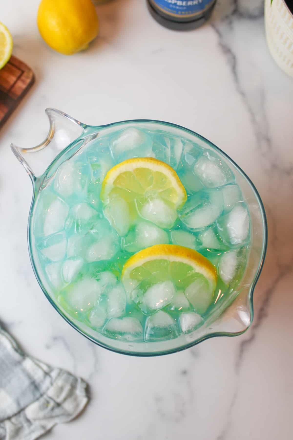 a pitcher full of blue lemonade and ice with lemon slices.